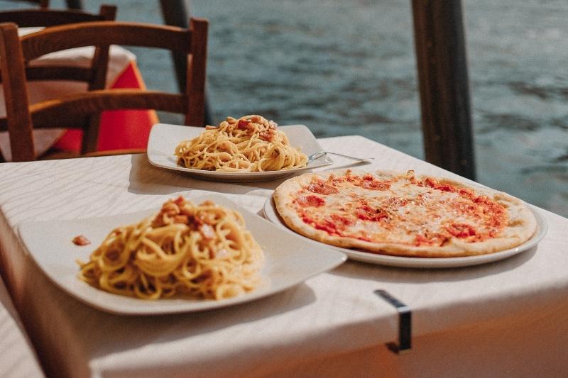 Image of pizza and two bowls and spaghetti on a table with white table cloth and wooden chairs.